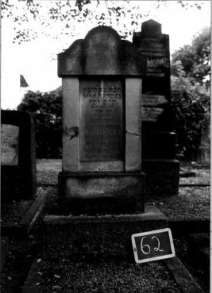 Grabstein 62 (Index: 100110282) Material: Sandstein (?) mit Marmorplatte Zustand: leicht beschädigt Maße HxBxT: 149x57x18 cm Beschreibung: Stele, getreppter Sockel, geschwungener Rundbogenaufsatz, eingetiefte Schriftplatte rechteckig: Grabstein 62