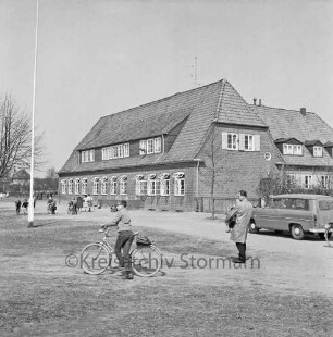 Volksschule: Hamburger Straße: vorn Junge mit Fahrrad: rechts Lehrer: dahinter Pkw: links Fahnenmast: hinten links weitere Schulkinder auf Schulhof