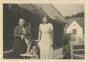 unbekannt: eine Frau, eine junge Frau, davor auf Klappstuhl Schäferhund: Rohlfshagen, Kupfermühle: Schäferkate, vor Traufseite: Plattenweg, links Bank, rechts Gartenstuhl, hinten Zaun, Gartentor, dahinter Atelier, Schuppen: im Hintergrund Wiese an Abhang