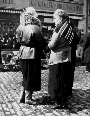 Gespräch auf dem Markt. Zwei gut gekleidete Frauen unterhalten sich. Im Hintergrund eine Buchhandlung
