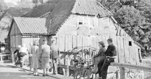 Grander Mühle mit Nebengebäuden: verfallenenes Gebäude und beschädigtes Mühlrad: vorn auf Holzbrücke mehrere Personen mit Fahrrädern: rechts Mühlenteich: hinten Bäume