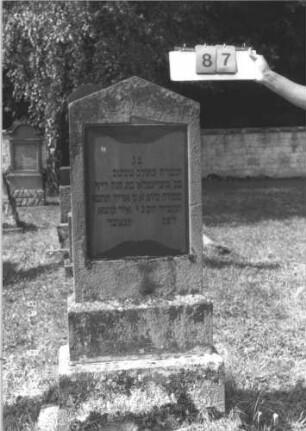 Grabstein 87 (Index: 99609723) Material: Sandstein ?, Platte Granit Zustand: stark verwittert, angeschlagen Maße HxBxT: 140x52x18 cm Beschreibung: Stele, Giebelabschluß, rechteckige Schriftplatte eingelassen, getreppter Sockel: Grabstein 87
