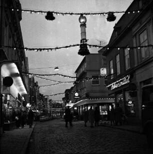 Mühlenstraße: Adventsbeleuchtung und erleuchtete Geschäfte: rechts Feinkostgeschäft Peters, dahinter Einmündung Hindenburgstraße: links Kaufhaus Nickel: dazwischen Personen: Abend