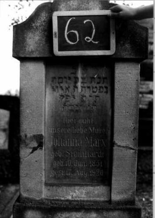 Grabstein 62 (Index: 100110282) Material: Sandstein (?) mit Marmorplatte Zustand: leicht beschädigt Maße HxBxT: 149x57x18 cm Beschreibung: Stele, getreppter Sockel, geschwungener Rundbogenaufsatz, eingetiefte Schriftplatte rechteckig: Grabstein 62 (Detailaufnahme)