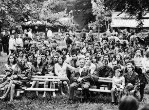 Parkfest : Besucher eines Festes im Park sitzen auf Bänken wie vor einer Bühne