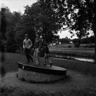 Kinderspielplatz: am Travewanderweg: drei Jungen auf Drehscheibe: Trave: am gegenüberliegenden Ufer Wiesen, Bungalow