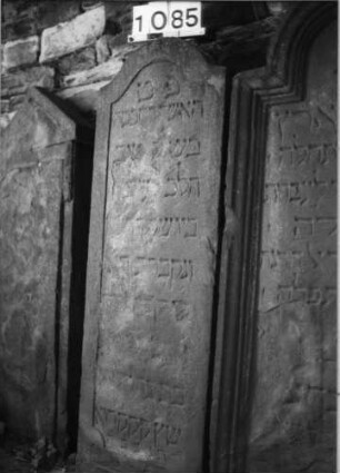 Grabstein 1085 (Index: 99630588) Material: Sandstein Zustand: stark verwittert mit Textverlust Maße HxBxT: 142x45x10 cm Beschreibung: Stele mit eingezogenem Segmentbogenabschluß, Schriftfeld eingetieft: Grabstein 1085