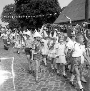 Kindervogelschießen: Umzug: geschmückte Straße: Schülerinnen und Schüler mit Papierhüten, Krönchen, Blumensträußen, Girlande