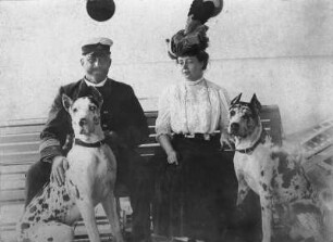 Kreuzfahrten der Hamburg-Amerika Linie, um 1911/1913. Kapitän, daneben eine Frau mit Hunden auf einer Bank an Deck eines Hochseepassagierdampfers, vermutlich der "Pennsylvania"