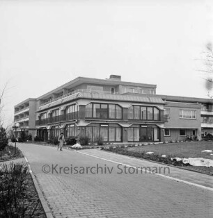 Seniorenwohnanlage Rosenhof I: Hoisdorfer Landstraße: Außenansicht des Gebäudekomplexes: vorn Zuwegung: darauf links Frau: rechts und links Grünanlage: Winter