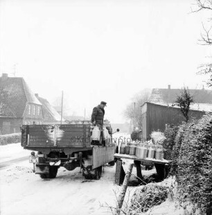 Dorfstraße: Lkw mit Milchkannen: rechts Milchbock: Mann beim Austauschen der Milchkannen: dahinter Schuppen: hinten Bauernhäuser: Winter