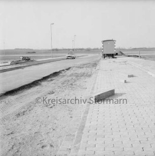 Gewerbegebiet West: Straßenbau: vorn unbefestigtes Gelände, gepflasterter Parkstreifen: hinten Straße, Fußwege, Straßenlampen, Bauwagen, Pkw und Baumaterial: dahinter Wiesen und Bäume
