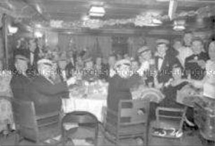Abendgesellschaft im geschmückten Festsaal des Hochseepassagierdampfers "Bremen", an Tischen sitzend, dem Fotografen zuprostend