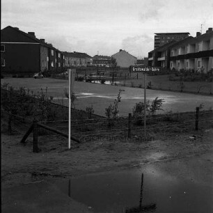 Greifswalder Kehre: vorne ein Parkplatz, dahinter Grünfläche mit kleinem Kinderspielplatz, an den Seiten Wohngebäude, rechts im Hintergrund das Rathausgebäude