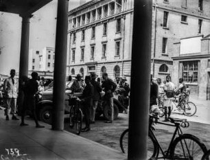 Straßenbild aus Salisbury : Blick auf eine Straße in Salisbury (heute: Harare). Mehrere Fahrzeuge, darunter mindestens ein Auto der Schomburgk-Expedition, sind vor einem Gebäude abgestellt. Darum herum eine Menschenmenge.