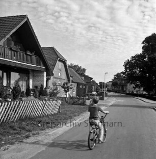 Dorfstraße: links und hinten rechts Einfamilien- und Bauernhäuser mit Vorgärten: vorn Mitte Kind mit Fahrrad