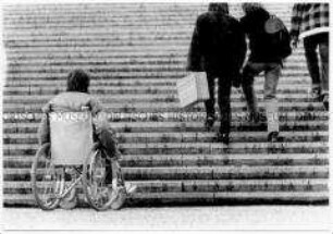 Mann im Rollstuhl am Fuß einer Steintreppe, auf der zwei Frauen nach oben gehen (Altersgruppe 18-21)