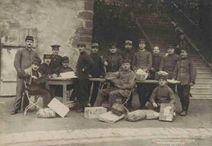 Fünfzehn Kriegsgefangene, teils stehend, teils sitzend an Tischen beim Verteilen von Paketen