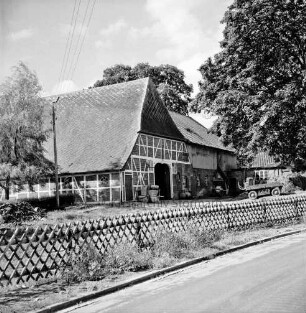 Fachwerkhaus eines Bauernhofes, dahinter ein angrenzendes Hofgebäude, rechts ein Anhänger