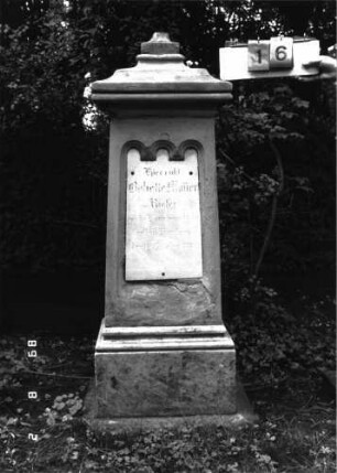 Grabstein 16 (Index: 100101734) Material: Sandstein, weiße Marmorplatte Zustand: unten abgeblättert, Aufsatz fehlt Maße HxBxT: 154x50x38 cm Beschreibung: konische Stele, profilierter Sockel, Abdeckplatte mit Aufsatz, Schriftplatte mit dreifachem Rundbogenfries Symbolik: gestelztes Rundbogenfries Steinmetz: Epple: Grabstein 16