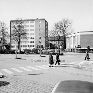 Erweiterungsbauten "Hochhaus" [später Gebäude B] und Kreistagssitzungssaal: dazwischen Hauptgebäude [später Gebäude A]: davor Parkplatz und Bäume: vorn Bahnhofsvorplatz mit Mommsenstraße: in der Mitte und rechts drei Personen auf Zebrastreifen