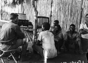 Werbeaufnahme für Telefunken : Blick auf eine Gruppe von sitzenden Männern, die sich vor einem Radio der Firma Telefunken versammelt haben.