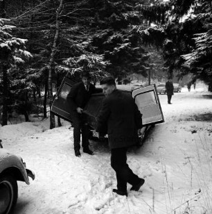 Polizei: Mord an der Hamburger Küchenhilfe Edeltraut Harloff am 26. November 1965: Oher Tannen: Leichenfundort zwischen Tannen und Totholz: Abtransport der Toten: in der Mitte Leichenwagen mit geöffneter Hecktür: davor zwei Bestatter mit Holzsarg: hinten rechts mehrere Polizeibeamte der Lübecker und der Hamburger Mordkommission auf Weg sowie parkende Pkws: hinten Tannen: Winter