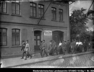 Arbeiter Wilhelm Lampe aus Wolfenbüttel, Johannisstr.1, als Sergeant des alten Braunschweigischen Infanterieregiments Enthält: Platte ist abgesplittert, Lampe geht mit einem Soldaten an einem Haus mit dem Schild: Richard Schulz Seifenfabrik, vorbei, dahinter: Soldaten mit Damen, paarweise gehend, Jugendliche