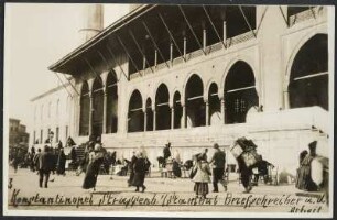 Blick auf eine Reihe von Briefeschreibern vor dem Eingang der Valide-Moschee in Istanbul, gestaltet mit Arkadenbögen und Vordach, im Vordergrund Passanten und Lastenträger