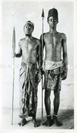 Porträt zweier Männer der Ndzimu mit Waffen vor neutralem Hintergrund