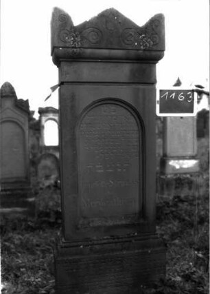 Grabstein 1163 (Index: 100104290) Material: Sandstein Zustand: gut erhalten, nur in Bodennähe verwittert Maße HxBxT: 176x56x22 cm Beschreibung: aufgesockelte Stele, Giebelaufsatz, Eckakroterien mit eingeritzten Voluten, Ranken und Blüte, eingetieftes Schriftfeld mit mehrfach profiliertem Rundbogenabschluss Symbolik: Voluten, Ranken, Blüte: Grabstein 1163