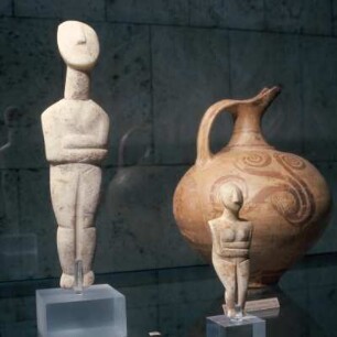 Stehendes weibliches Idol : München, Antikensammlung. Weibliches Idol, frühe Spedos-Art, frühkykladisch II. Rechts: Weibliches Idol mit verschränkten Armen (Spedos-Art), Inv.Nr. 263. Im Hintergrund rechts: Kanne, Mykenisch, um 1400 v. Chr., Ton, bemalt