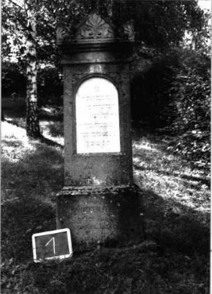 Grabstein 1 (Index: 100100923) Material: Kalkstein mit weißer Marmorplatte Zustand: leicht verwittert Maße HxBxT: 162x45x19 cm Beschreibung: Stele, Sockel, Giebel mit Eckakroteren, eingetiefte Schrifttafel mit Rundbogen Symbolik: Plametten im Giebel: Grabstein 1