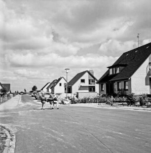 Neubausiedlung: rechts Einfamilienhäuser, einige vor Fertigstellung: davor Sandhaufen und Pkws: links drei Schulkinder: vorn Straße