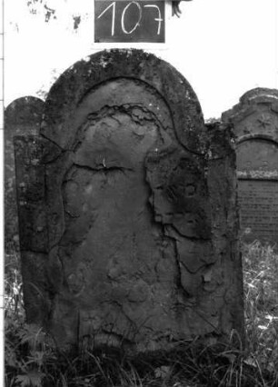 Grabstein 107 (Index: 100103519) Material: Sandstein Zustand: stark verwittert, abgeblättert, fast vollständiger Textverlust Maße HxBxT: 106x64x5 cm Beschreibung: Stele, eingezogener Rundbogenabschluss, Schriftfeld eingetieft mit Konvexbogenabschluss: Grabstein 107