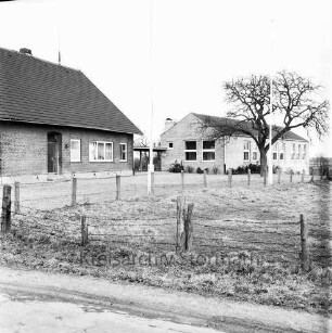 Schmachthagen: Dorfstraße / Schulsteig: Volksschule: links altes Gebäude, rechts Erweiterungsbau: davor Schulhof mit Baum und Fahnenmasten: im Vordergrund Wiese mit Zaun
