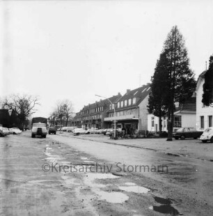 Eilbergweg: vorn mit Wasser gefüllte Schlaglöcher in Fahrbahn: rechts Baum: dahinter Wohn- und Geschäftsblocks: davor Parkplätze mit Pkws: hinten links Lkw und Pkws: dahinter Bäume