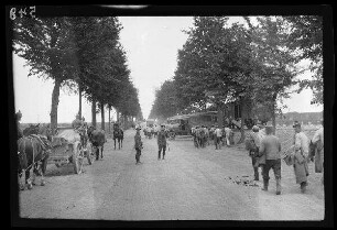 "An der Straße Cambrai (Dep. Nord, Frankreich) - Bapaume (Dep. Pas-de-Calais, Frankreich)": Fuhrwerkkolonne und Lastkraftwagen auf einer Allee, am Rand ein Eisenbahnwaggon (Rückansicht)