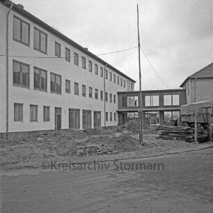 Ahrensburg: Hermann-Löns-Straße: Erweiterungsbau: vor Fertigstellung: hinten Bildmitte Verbindungstrakt, rechts Altbau: auf Vorplatz Baustelle, Erdhaufen, Gerüstteile, Mauersteine, Lkw, provisorischer Strommast, Arbeiter