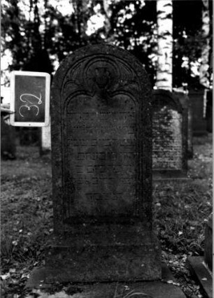 Grabstein 32 (Index: 100111070) Material: Sandstein Zustand: leicht verwittert Maße HxBxT: 116x48x16 cm Beschreibung: Stele, Sockel, Rundbogenabschluß, Schriftfeld eingetieft mit Kleeblattbogenabschluß Symbolik: Levitenkanne, Maßwerk Besonderheit: Kindergrab: Grabstein 32