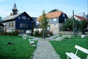 Dorfplatz mit Grünfläche und gepflastertem Weg, vielen Bänken
