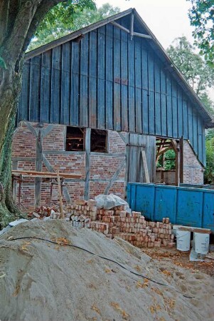 Dorfplatz (2): Fachwerkscheune: Sanierung: Giebelseite: Gefache mit neuer Ausmauerung, Scheunentor, oben Blechverkleidung, Giebelzier: davor Stapel mit Backsteinen, Sandhaufen, Schuttcontainer: Juli 1994