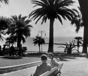 Italien. Alassio an der Ligurischen Küste. Aufnahme der Strandpromenade mit Blick auf gleichnamige Bucht. Touristen sitzen auf einer Bank