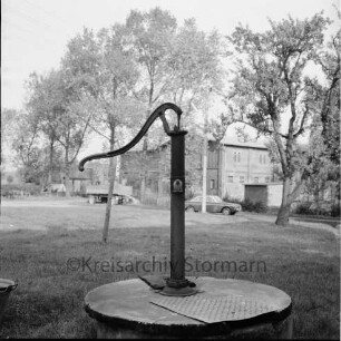 Dorfplatz mit Bäumen: vorn Brunnen mit Pumpe: im Hintergrund Bauerngehöft: davor Pkw und landwirtschaftlicher Anhänger