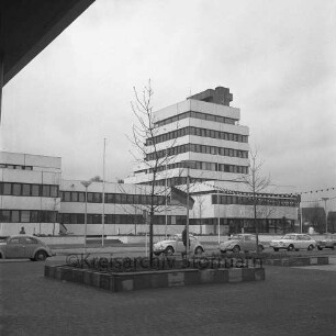 Rathaus: davor Rathausvorplatz mit Springbrunnen, Fahnenmasten, Deutschlandfahne auf Halbmast: Straße Reeshoop [später Manfred-Samusch-Straße], parkende Autos, Fußgänger, Straßenlampen: vorne auf Rathausplatz zwei Grüninseln mit Sitzkanten und Bäumen, Lichterketten: links oben überhängendes Dach von Arkaden der Randbebauung