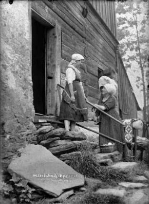 Steinbach im Erzgebirge. Zwei junge Frauen in bäuerlicher Bekleidung vor einem Blockbohlenhaus, vom Klöppeln zurückkehrend