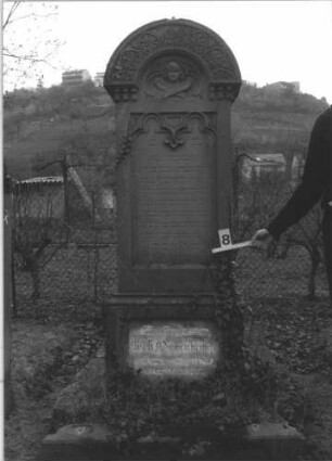Grabstein 8 (Index: 99805015) Material: Sandstein Zustand: deutsche Inschrift im Sockel verwittert Maße HxBxT: 195x68x28 cm Beschreibung: Stele auf Doppelsockel mit Rundbogen, Inschriftennische rechteckig mit zwei Kleeblattbögen als Verzierung, Inschriftennische im Sockel querrechteckig Symbolik: Engel im Rundbogen, Blattornamentik am Rundbogen: Grabstein 8