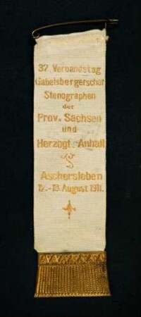 Bandabzeichen 32. Verbandstag Gabelsbergerscher Stenographen der Provinz Sachsen und des Herzogtums Anhalt in Aschersleben 1911 (mit Broschennadel) : Medaillensammlung Stenografische Sammlung. Bandabzeichen 32. Verbandstag Gabelsbergerscher Stenographen der Provinz Sachsen und des Herzogtums Anhalt in Aschersleben 1911 (perlmuttfarbenes Band mit goldener Aufschrift "32. Verbandstag Gabelsbergerscher Stenographen der Prov. Sachsen und Herzogt. Anhalt Aschersleben 12.-13. August 1911.", mit Broschennadel; rückseitig Herstellerstempel: Hallesche Fahnenfabrik Halle, S. Leipzigerstr. 72). Stoff; Maße (ohne Nadel) 100 x 27 mm (1911; Hallesche Fahnenfabrik). Dresden: SLUB R c 0418