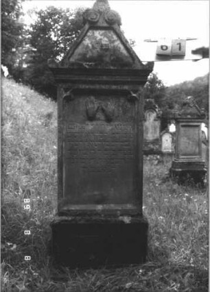 Grabstein 61 (Index: 99799529) Material: Sandstein Zustand: stark verwittert, versalzen Maße HxBxT: 118x62x22 cm Beschreibung: Stele, Abdeckplatte, Giebelaufsatz, Giebel abgeschlossen mit Palmette u. Voluten, Seitenakrotere, Schriftfeld eingetieft, Sockel Symbolik: segnende Hände: Grabstein 61