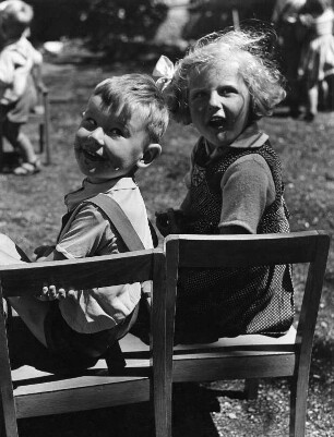 Kinderbilder. Kleines Mädchen und kleiner Junge, auf Stühlen sitzend und sich umwendend (vielleicht in der Vorschule)